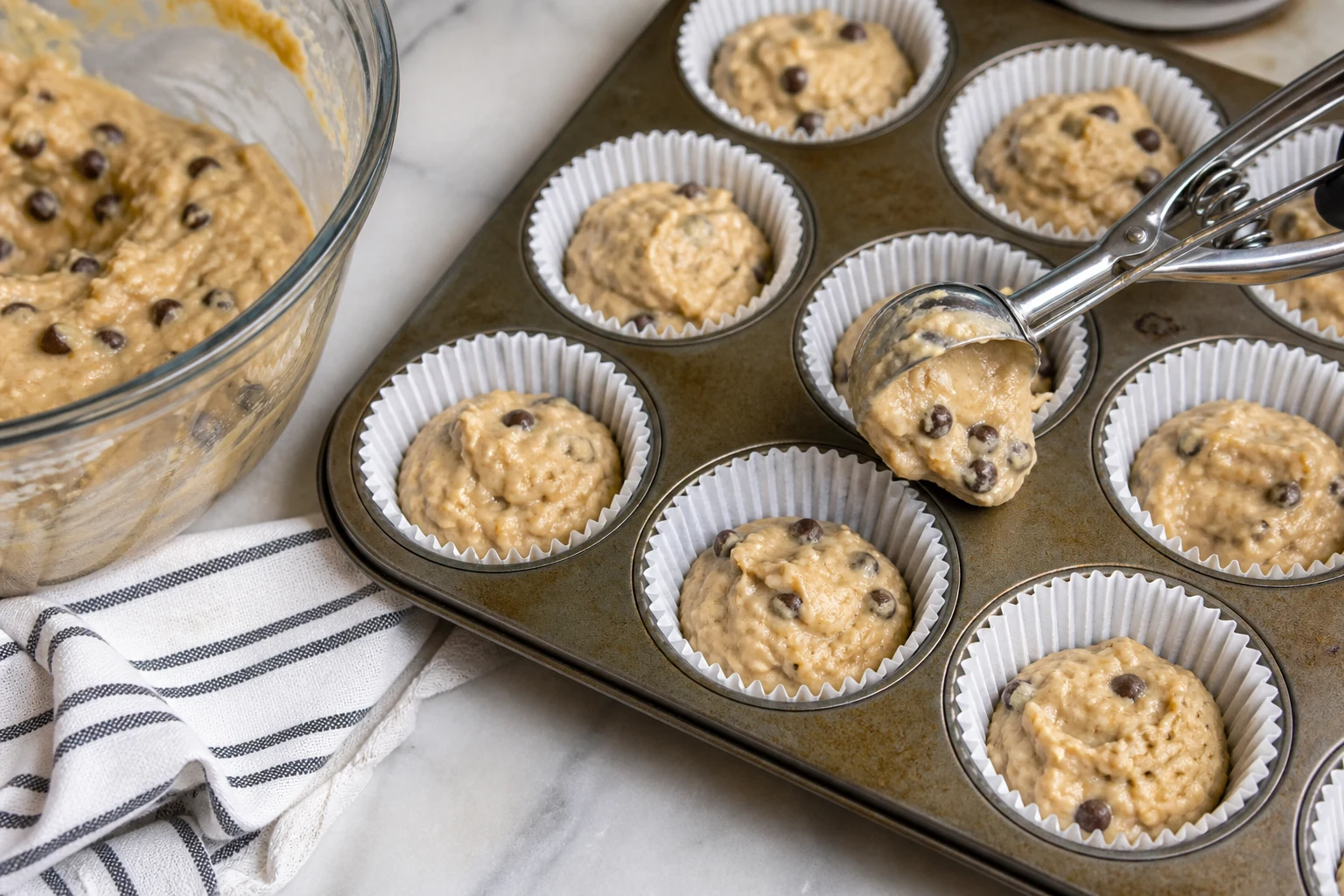 Thick banana chocolate-chip batter being portioned into lined muffin cups before a hot-start bake.