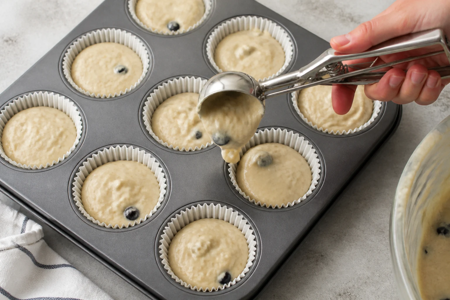 Thick pale Greek-yogurt muffin batter being portioned evenly into a lined 12-cup pan.
