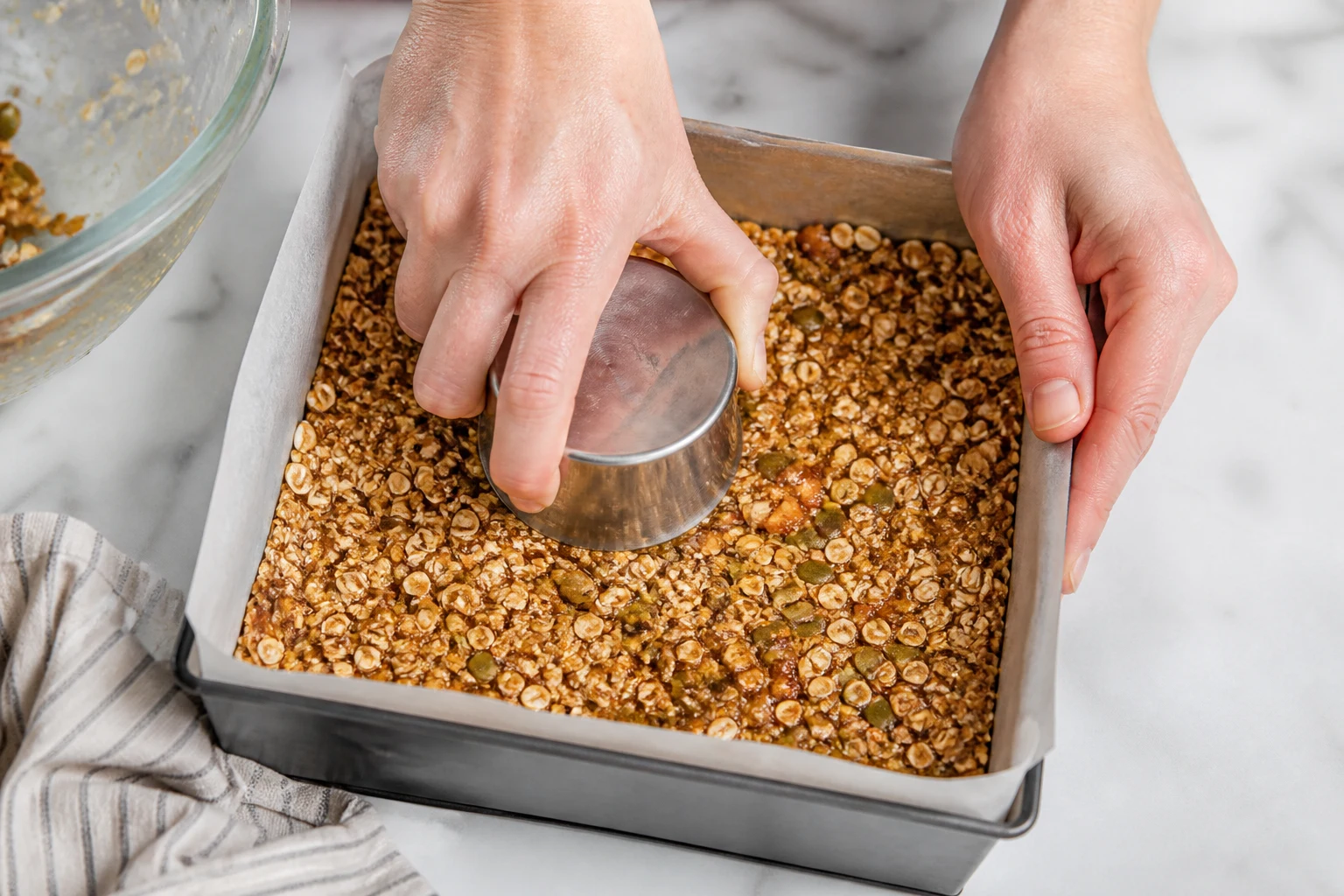 Warm coated granola-bar mixture being pressed very firmly into a parchment-lined square pan.