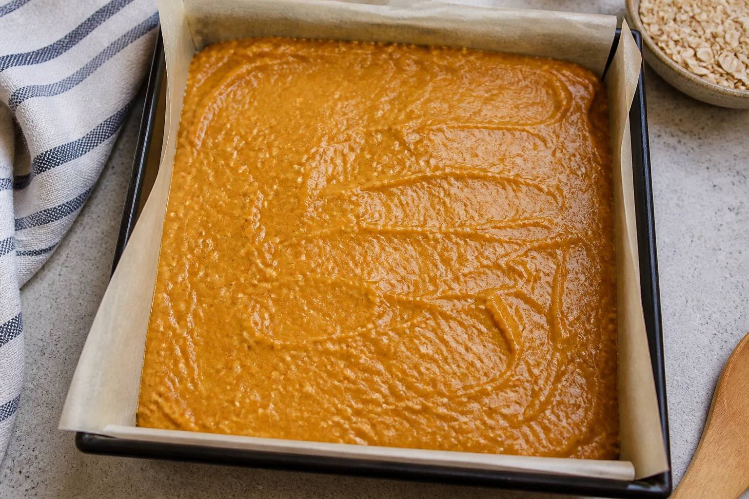 Smooth pumpkin-oat batter spread in a parchment-lined square pan before baking.