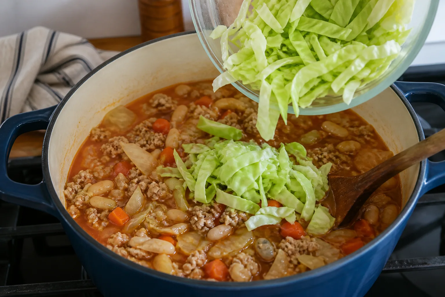 Second portion of green cabbage ribbons being added to turkey and white bean soup in a Dutch oven