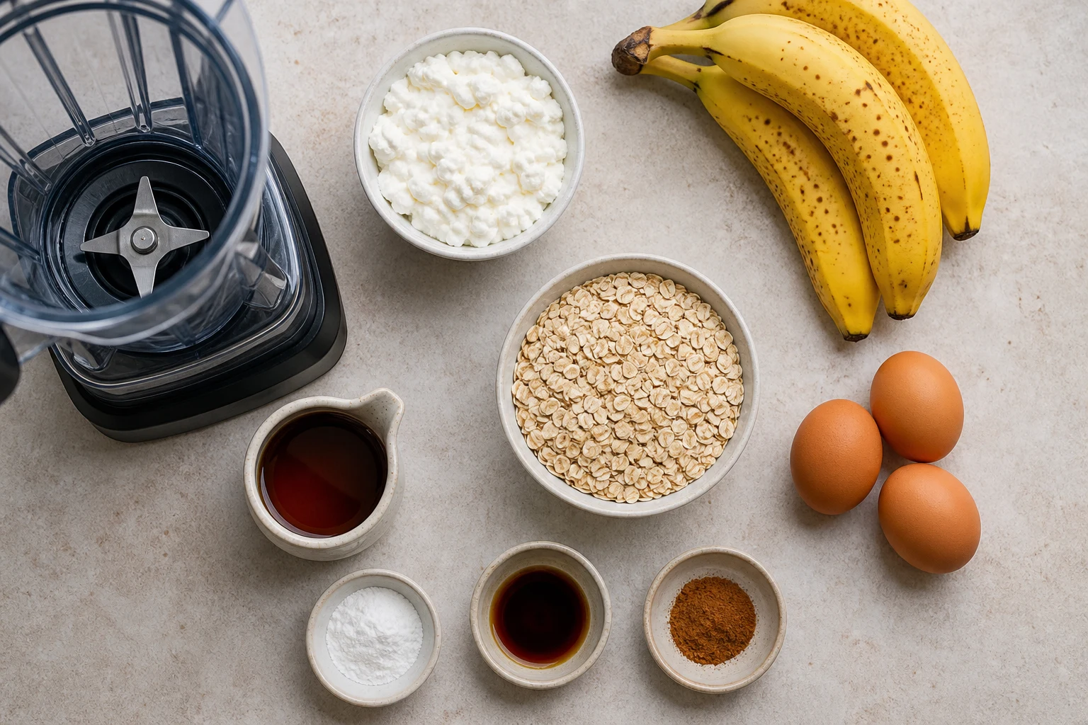 Rolled oats, cottage cheese, eggs, ripe bananas, maple syrup, baking powder, vanilla and cinnamon beside a blender.