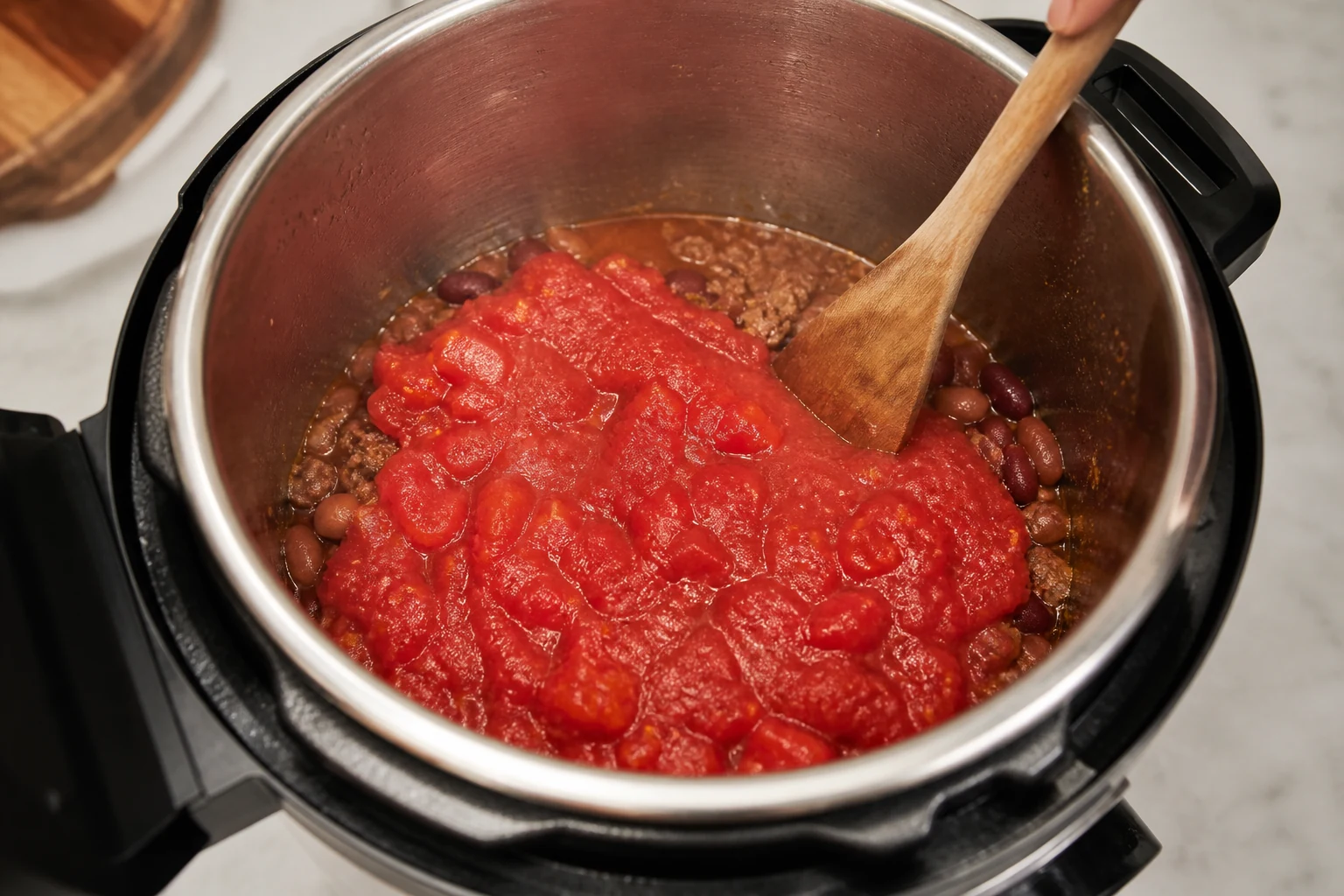 Electric pressure-cooker insert after deglazing, browned beef and beans below with crushed tomatoes layered visibly on top and not stirred into the base.