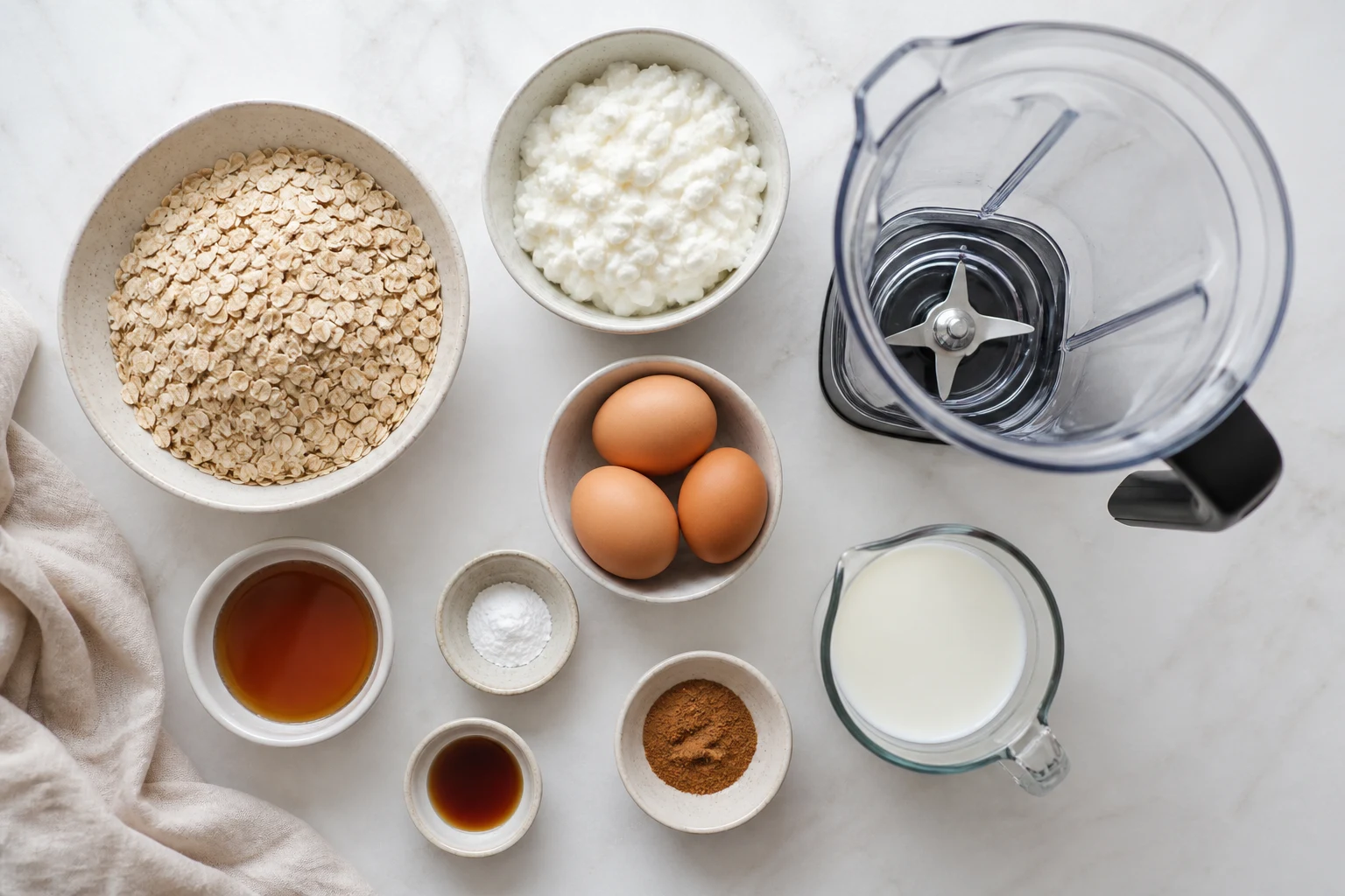 Rolled oats, cottage cheese, eggs, maple syrup, baking powder, vanilla, cinnamon and milk beside a blender.