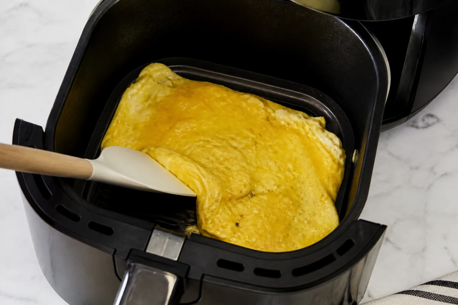 Partially set scrambled eggs being folded from the edges with a silicone spatula inside a small air-fryer pan.